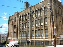 A four-story brown brick school building sits behind a black metal fence and crisscrossing overhead telephone and electrical cables.