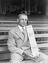 Man in double breasted suit, hair parted down the middle, sitting on a long bench in a sports stadium, posing with a cricket bat, held vertical and supported on his thigh.