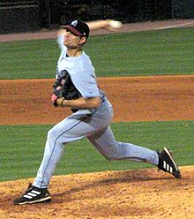 Ein Mann in einer grauen Baseballuniform und einer schwarzen Kappe bereitet sich darauf vor, einen Baseball aus seiner rechten Hand zu werfen, während er auf dem Hügel des Werfers steht.