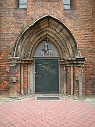 Portal romańsko-gotycki z gotycką wimpergą, fragmentem tympanonu i ekspresjonistyczną stolarką z lat 30. XX w.