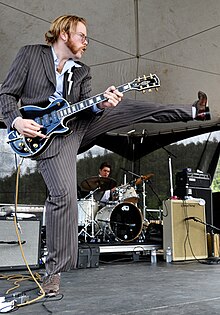 A photograph of guitarist Kid Andersen kicking a leg in the air while playing guitar.