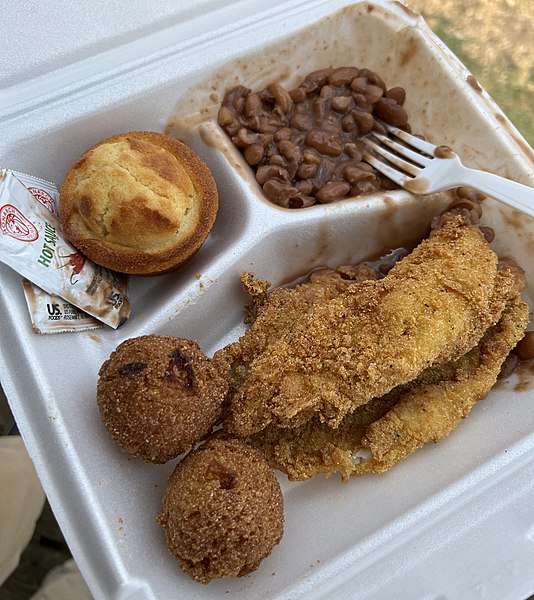 A typical meal of fried catfish filets paired with hushpuppies, pinto beans, cornbread, and hot sauce.