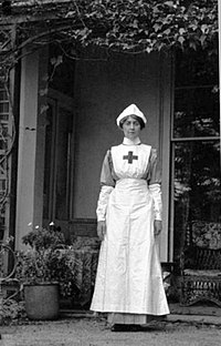 A black and white photograph of Agatha Christie in the uniform she wore as a nurse and dispenser when working as a member of the Voluntary Aid Detachment. The uniform consists of a white hat and a white apron with a cross on the front.