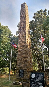 Tall field stone column memorial flanked by United States and Confederate States of America flags surrounded by tall, leafy green trees.