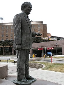 A bronze statue of a late middle-aged man holding up his hand at waist level.