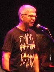 A gray-haired man with glasses and a black shirt standing in front of a microphone