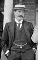 Schwarzweißfoto eines schlanken Mannes mit Schnauzbart, der einen dunklen Dreiteiler und einen Hut trägt