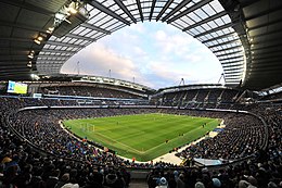 Photo panoramique du stade de Manchester City. Les spectateurs sont tous assis, le terrain est au centre de l'image. Le ciel est dégagé avec quelques nuages. Le match est en cours. Un panneau d'affichage annonce que le score est de zéro à zéro.
