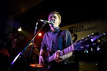 Peter Brewis of the rock band Field Music performing on a stage, playing a guitar and singing into a microphone, as other musicians perform in the darkened background behind him.