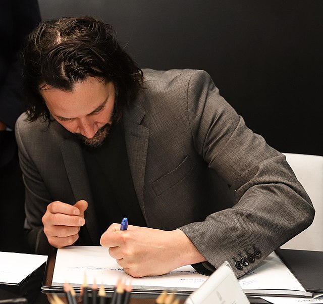 Image shows Keanu Reeves writing in a notebook with his left hand.
