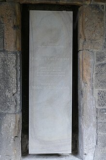 Grauer, rechteckiger Stein mit Inschrift hochkant in eine Wand eingelassen