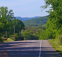 Eine vierspurige Autobahn führt einen Hügel hinab und verschwindet aus dem Blickfeld.  Im Hintergrund sind große, bewaldete Berge.