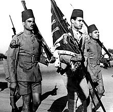 Photographie de trois hommes en uniforme militaire. L'homme au centre tient un drapeau tandis que ses voisins portent un fusil.