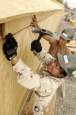 Two people standing on a scaffold wearing hard hats and protective gloves while hammering a nail into the side of a wooden building