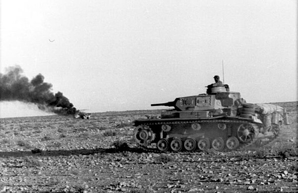 Panzer III advances past a burning vehicle in the desert, April 1941