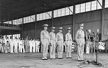 Макартур стоит внутри униформа у четырех микрофонов на стендах. Позади него по очереди стоят четверо мужчин в военной форме. Там видна большая толпа хорошо одетых мужчин, женщин и детей в юбках, костюмах и униформе. 