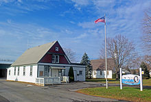 Til venstre en enetages træbygning med et højt tagtak.  Jordoverfladen er hvid med grøn kant;  det øverste niveau en mørk rødbrun, også med grøn kant.  I midten er en flagstang med det amerikanske flag;  bag den er en hvid kirke delvis skjult af et bare træ.  Til højre er der et blåt og hvidt skilt med "Clermont" fremtrædende på