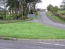 This image depicts a quiet rural crossroads with a well-maintained, grassy area in the foreground, the road curve slightly as it continues into the distance line with trees and fields on either side. A few buildings are visible further down the road with one prominent brick house surrounded by trees near the left side of the image, their traffic science present, including a sign on the grassy area in the middle, but they are not fully visible from this angle. The overall scene is serene with minimal traffic and a peaceful countryside ambience.