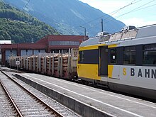Ein im fahrplanmäßigem Personenverkehr verkehrender MBS-Triebwagen mit angehängten Holztransportwagensteht im Bahnhof Schruns zur Abfahrt nach Bludenz bereit (2015); im Hintergrund die Diesellok V 10.017, mit welcher der Güterzug bereitgestellt wurde.