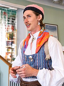 Bust of a tall, slender, white person with a painter's brush mustache wearing Bäckadräkten and speaking