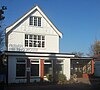 A converted house behind another house, whose side wall partly obscures the meeting house. A car is parked in front of the ground floor, which has three large windows with brickwork between them. The upper storey is painted white and bears the words "FRIENDS MEETING HOUSE".