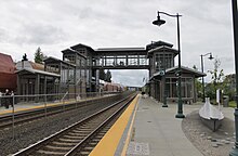 Жп гара с две коловози и две платформи от всяка страна, свързани с надземен стъклен мост