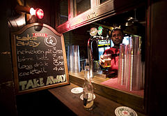 A bartender offers a beer through a bar window at night, with Bar signs at side, Rome, Italy