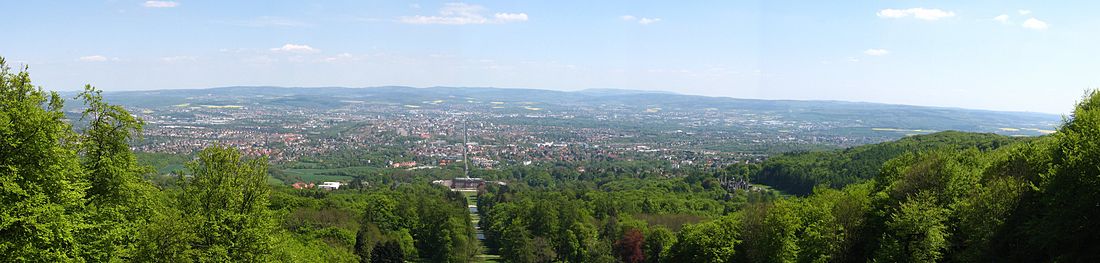 Blick vom Fuß des Herkules über den Bergpark auf Kassel: In der Bildmitte ist die zentrale Parkachse mit Schloss Wilhelmshöhe zu sehen, die ihre Verlängerung in der Wilhelmshöher Allee findet. Rechts im Bild ist auf halber Höhe die Löwenburg zu erkennen.