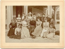 Schwarz-Weiß-Foto einer Gruppe von 16 Frauen vor einem Gebäude, teils mit Hüten, die sich in zeitgenössischen, festlichen langen Kleidern oder Blusen mit langen Röcken, zu einem Gruppenfoto aufgestellt haben. Die hintere Reihe von neun Frauen steht, die vorderen Personen sitzen auf Stühlen oder auf dem Boden.