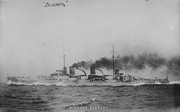 A light gray warship steaming at high speed; thick black smoke pours out of the two funnels.