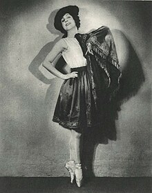 A white woman dancer, posing in toe shoes, one hand on a hip, wearing a hat, a shawl, and a short dark skirt