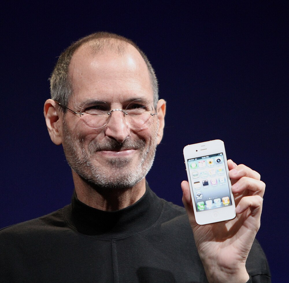 Shoulder-high portrait of smiling man in his fifties wearing a black turtle neck shirt with a day-old beard holding a phone facing the viewer in his left hand