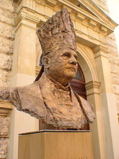 Farbfotografie von der Bronzebüste eines Mannes mit liturgischer Kopfbedeckung. Im Hintergrund ist ein Säuleneingang mit Giebel von einem hellen Gebäude zu sehen.