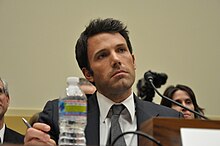 Ben Affleck, holding a pen and sitting behind a microphone, looks ahead while offering testimony