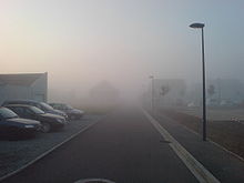 Un épais brouillard matinale envahi une route bordée de parkings.