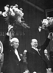 Two smiling men raise bouquets of flowers over their heads.