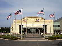 Above the doorway of a large, relatively plain rectangular structure with a short dome are the words "George Bush Library". In front of the building is a circular courtyard with a water fountain; eight American flags are positioned evenly around the circle.