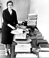 Black-and-white photo of Kelsey standing beside a table laden with files; grasping eyeglasses and an open book, she looks to the camera and seems about to speak