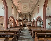 L'interno di un edificio rettangolare, pieno di panchine ordinate su ciascun lato di una navata centrale.  I lati della stanza hanno colonnati.  Il tutto è decorato in modo elegante, ma essenziale.