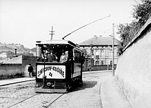 Otevřený tramvajový vůz jedoucí po vydlážděné ulici obehnané kamennými zdmi. Na voze je patrná ke krajnici vychýlená napájecí kladka.