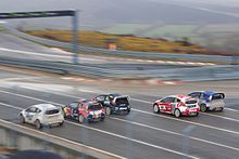Fotografia de cinco carros de rallycross, vistos de trás, partindo, alinhados, em cores diferentes.