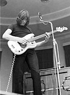 Ein monochromes Bild von Waters, der Bassgitarre spielt.Er hat schulterlanges Haar, schwarze Kleidung und steht vor einem Mikrofon.