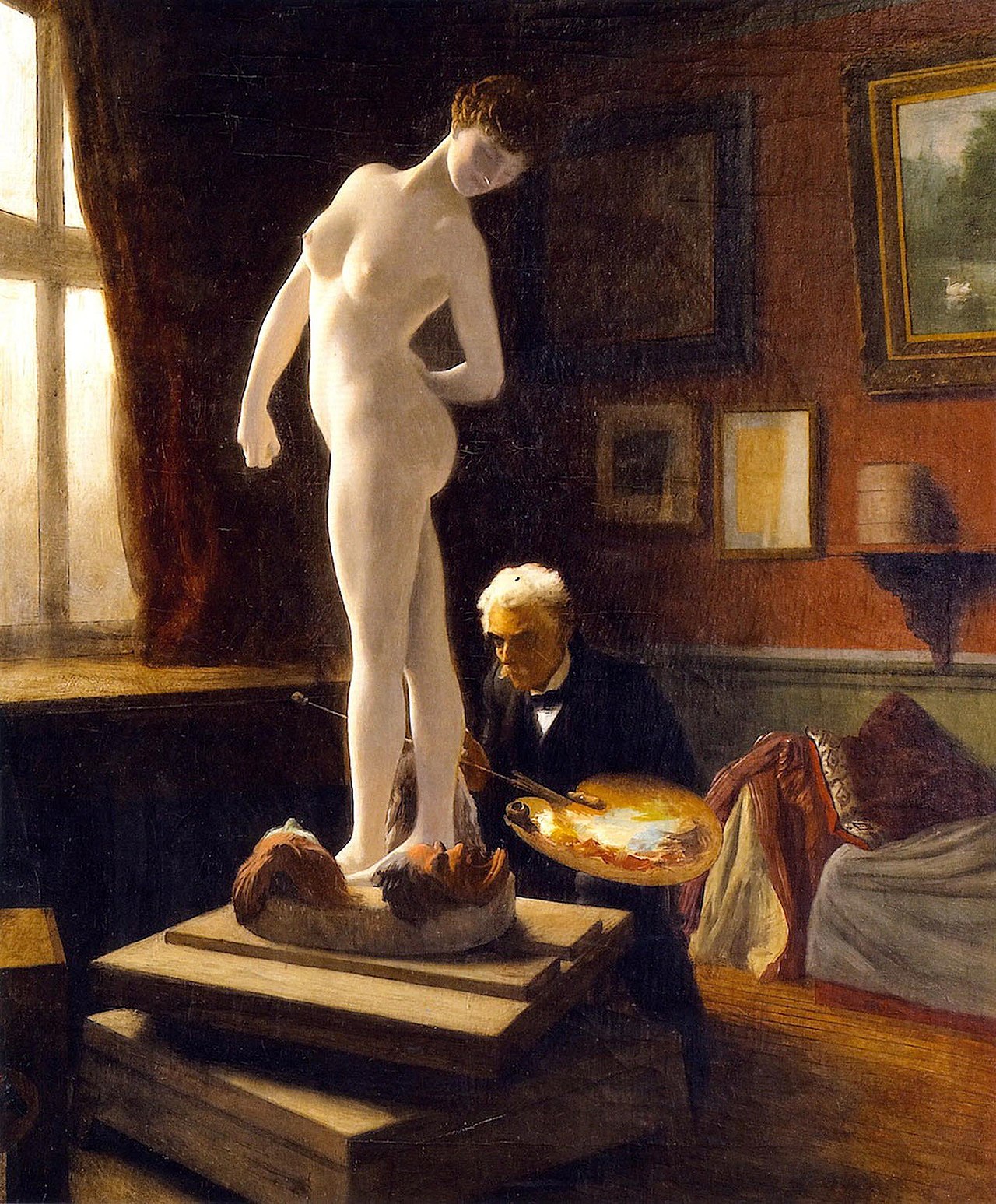 A male artist kneels at the feet of a female nude sculpture painting elements on the base of statue in a room with a couch, red walls and paintings on the walls as light streams in from a open window on the left.