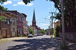 A view down a city street with a group of rowhouses of differing heights and colors on the left. A tall church spire is visible in the distance.