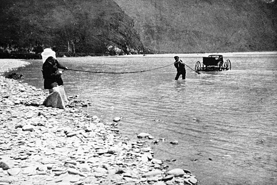A four-wheeled carriage in a calm river is pulled ashore with a rope by two men - one near the carriage with the rope over his shoulder, and the other pulling the rope from the gravel river bank.