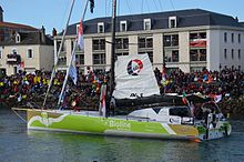 Bateau vert et marron progressant au moteur dans le chenal. O'Coineen est à l'avant, tenant un tambourin. Foule sur le quai.