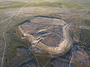 Aerial view of a massive hill, on which the ruins of walls and buildings can be seen, surrounded by wilderness