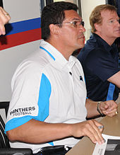 Sitting at a table, a man wearing a white Panthers shirt and glasses is signing an autograph.