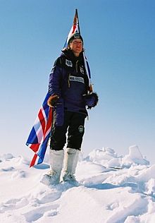 Eine Gestalt, die in dunkler Kleidung und weißen Stiefeln auf Schnee steht, trägt eine britische Flagge.