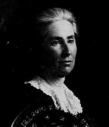 An older white woman, photographed mostly in shadows, with grey hair and wearing a white lace collar or kerchief with a dark garment